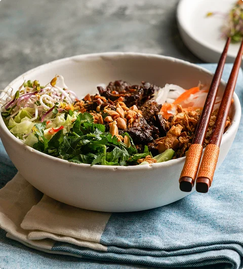 Een Vietnamese noedelkom met gegrild vlees, verse kruiden, groenten, pinda’s en eetstokjes.